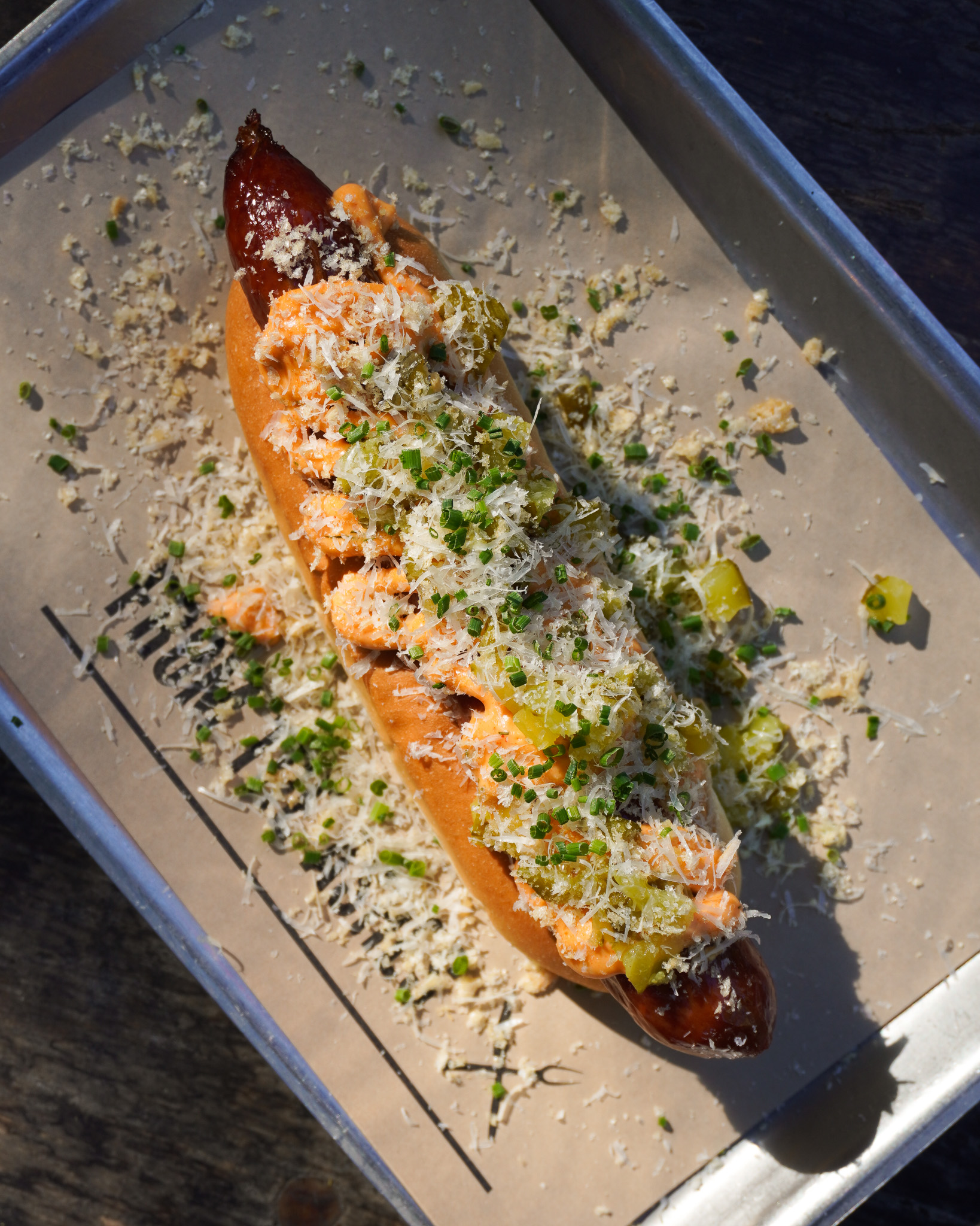 A hot dog lies on a tray with From the Ashes greaseproof paper. The hot dog is topped with cheese sauce, parmesan and chopped pickles