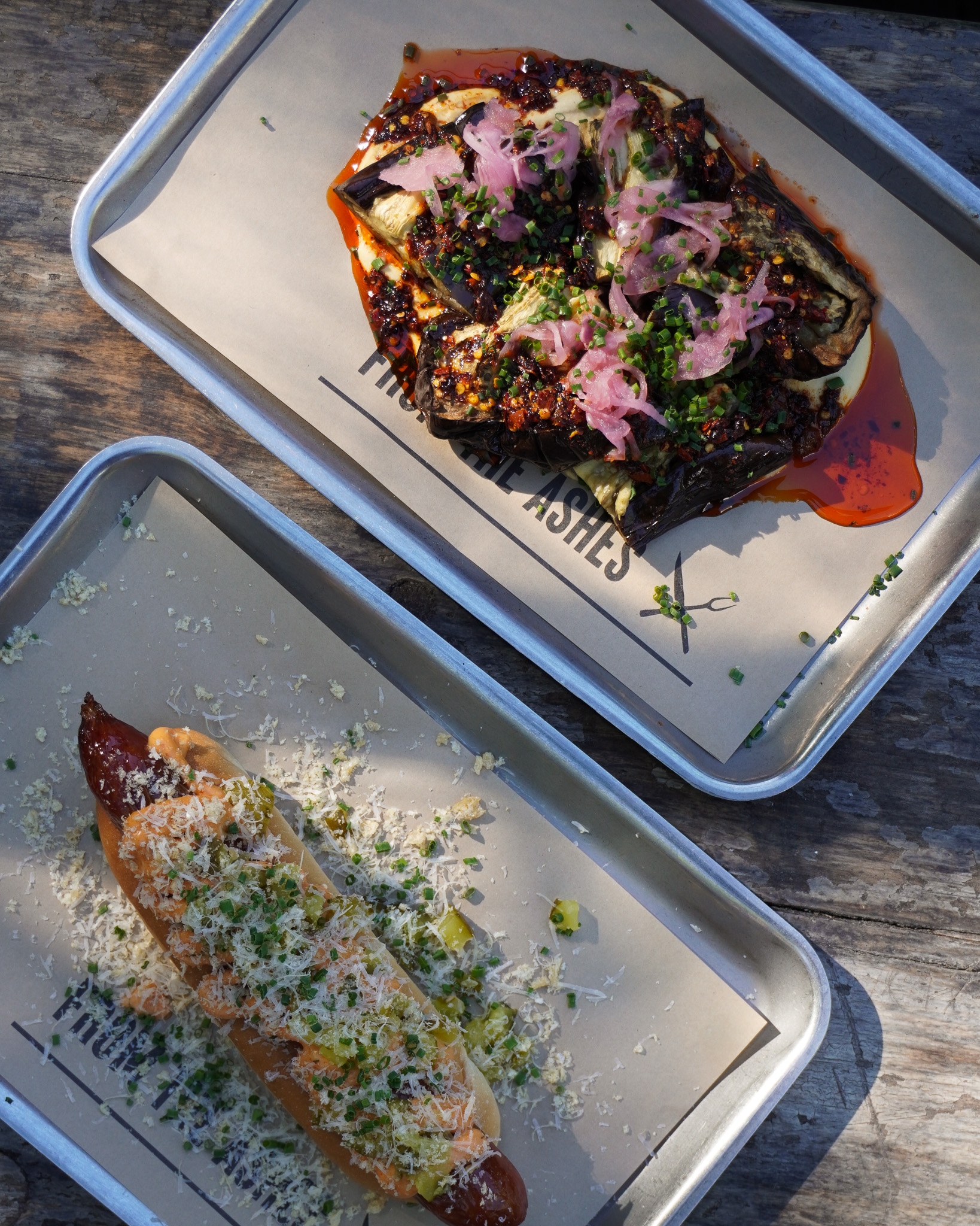 Two trays next to each other on a table viewed from above. One tray has roasted aubergine and the other has a hot dog on it