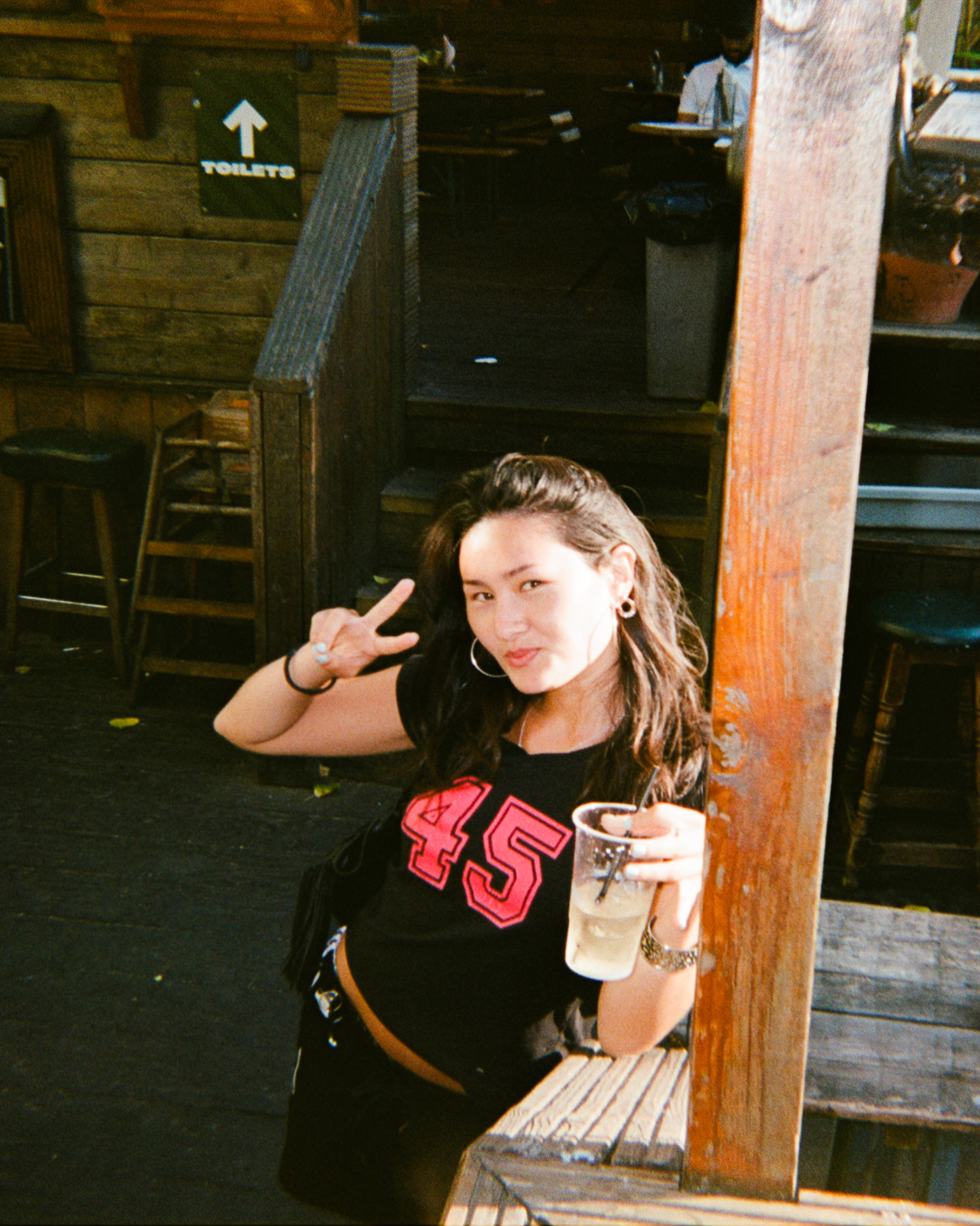 Photo of a woman making a peace sign with her right hand and a drink in her other hand leaning against a pillar in the beer garden